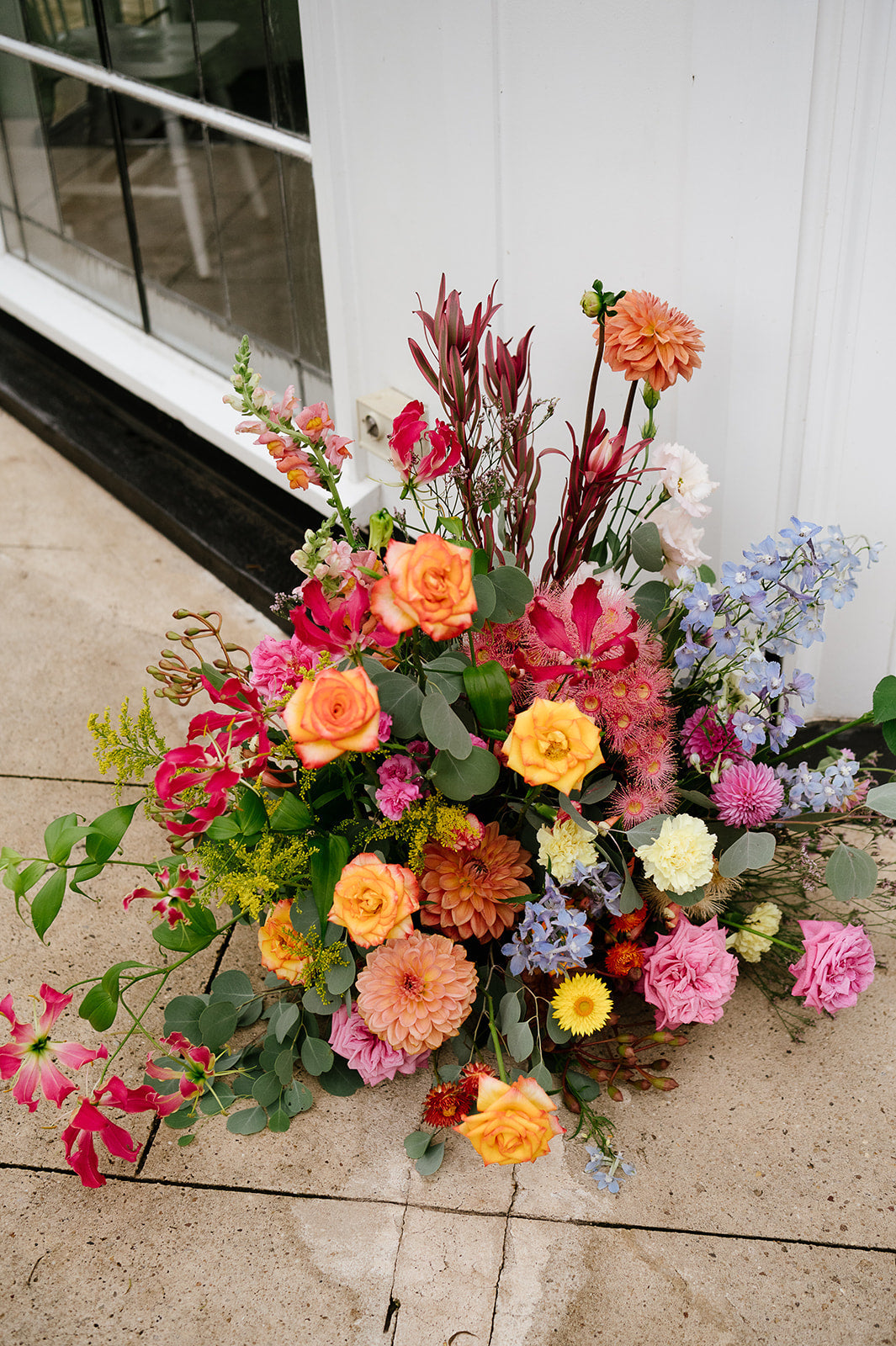 Large low arrangement of orange and yellow dahlias, pink blooms and blue delphinium on the paving beside the white gazebo at Kalinya Estate, by Lime Tree Bower Florist