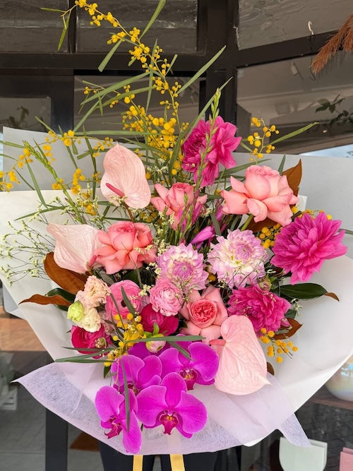 A bouquet of pink flowers including roses, orchids, and other seasonal flowers wrapped in paper.