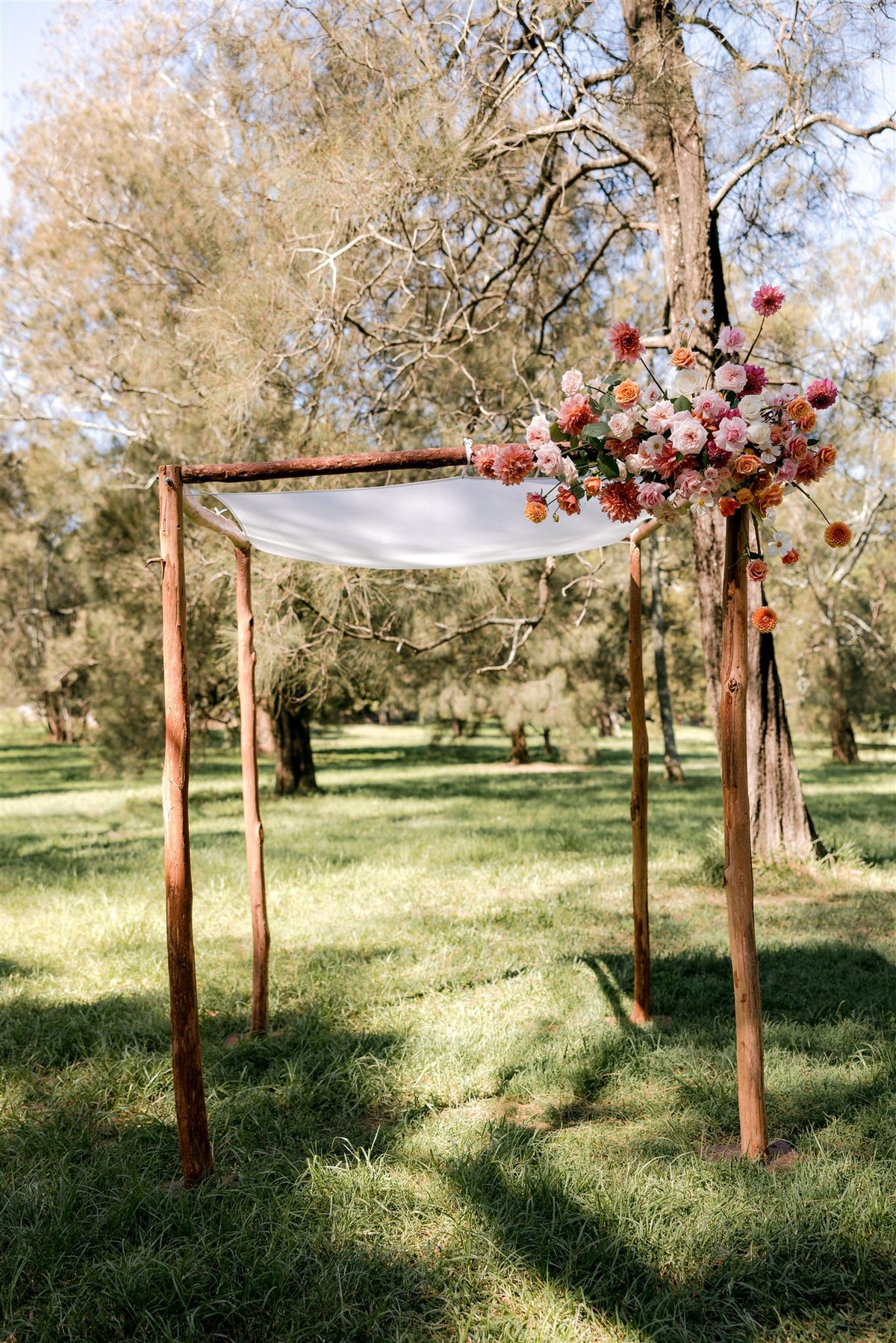 Timber chuppah with a white canopy and a single corner cluster of coral, peach and pink flowers under the trees in Centennial Park