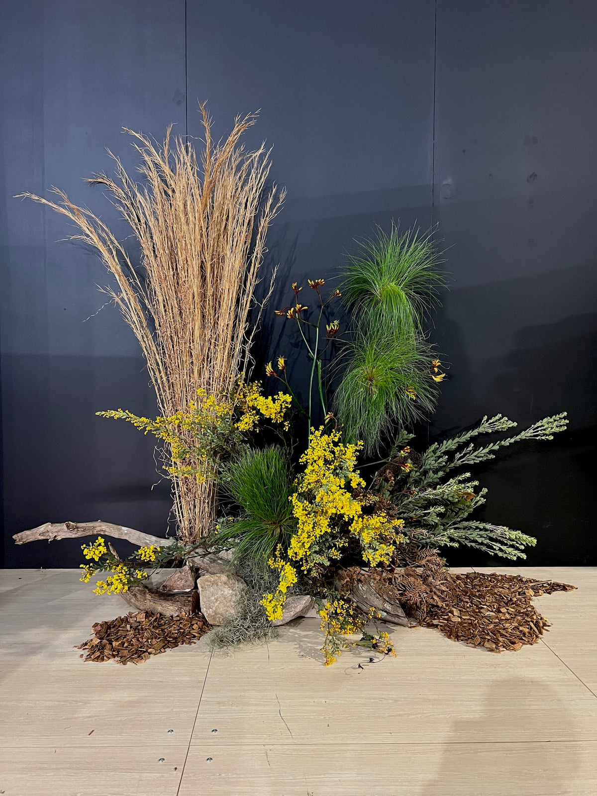 Decorative floral installation arrangement of grasses, foliage, native flowers and dried flowers against a dark backdrop on the floor for a sydney corporate event