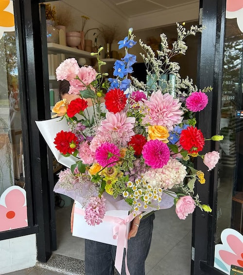 Bouquet of colourful, fresh flowers with dahlias, delphinium, chamomile daisy, roses, lisianthus, hydrangea, gerberas, freesias, wrapped and boxed, held by a person in front of a Sydney flower shop entrance.