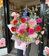 Bouquet of colourful, fresh flowers with dahlias, delphinium, chamomile daisy, roses, lisianthus, hydrangea, gerberas, freesias, wrapped and boxed, held by a person in front of a Sydney flower shop entrance.