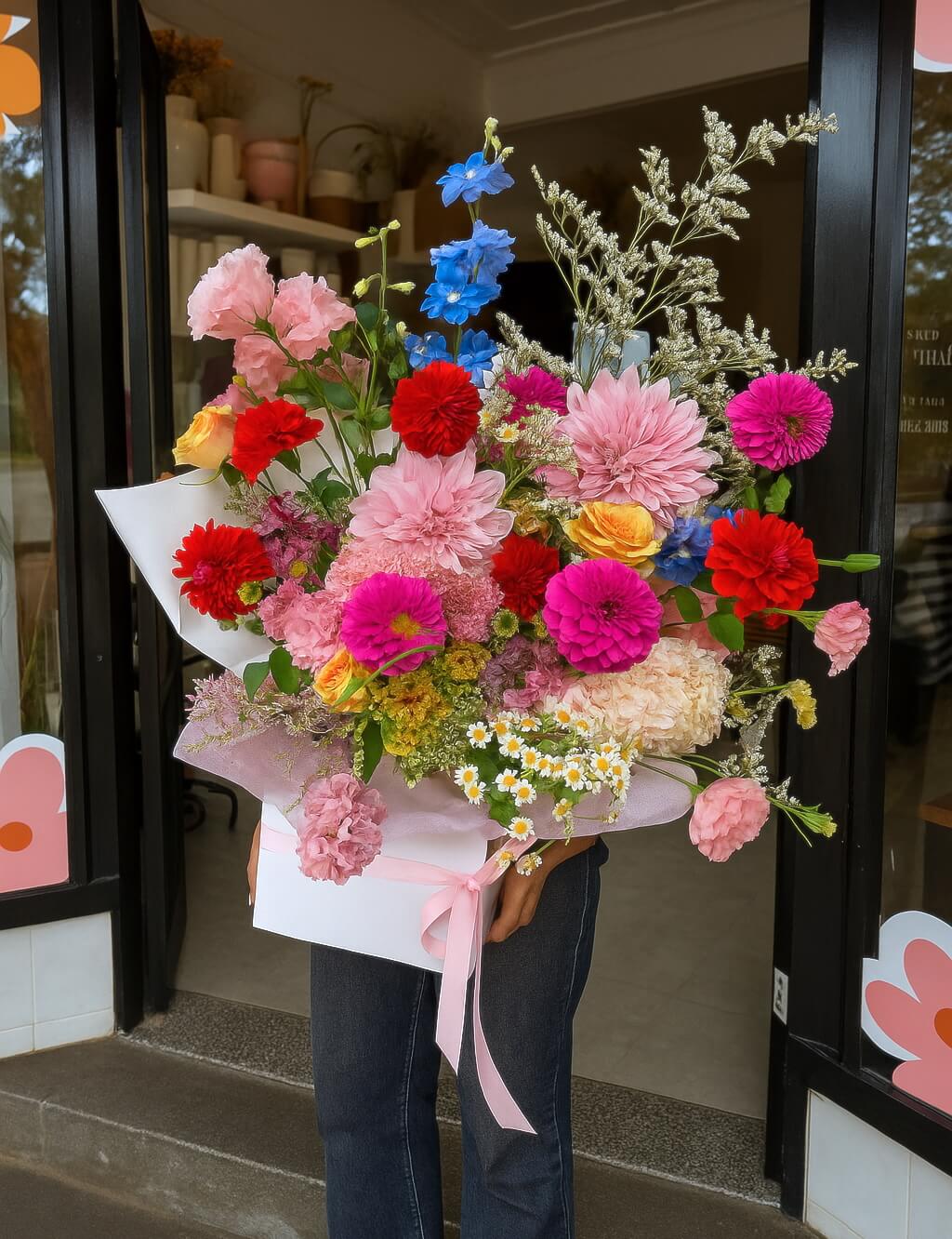 Bouquet of colourful, fresh flowers with dahlias, delphinium, chamomile daisy, roses, lisianthus, hydrangea, gerberas, freesias, wrapped and boxed, held by a person in front of a Sydney flower shop entrance.