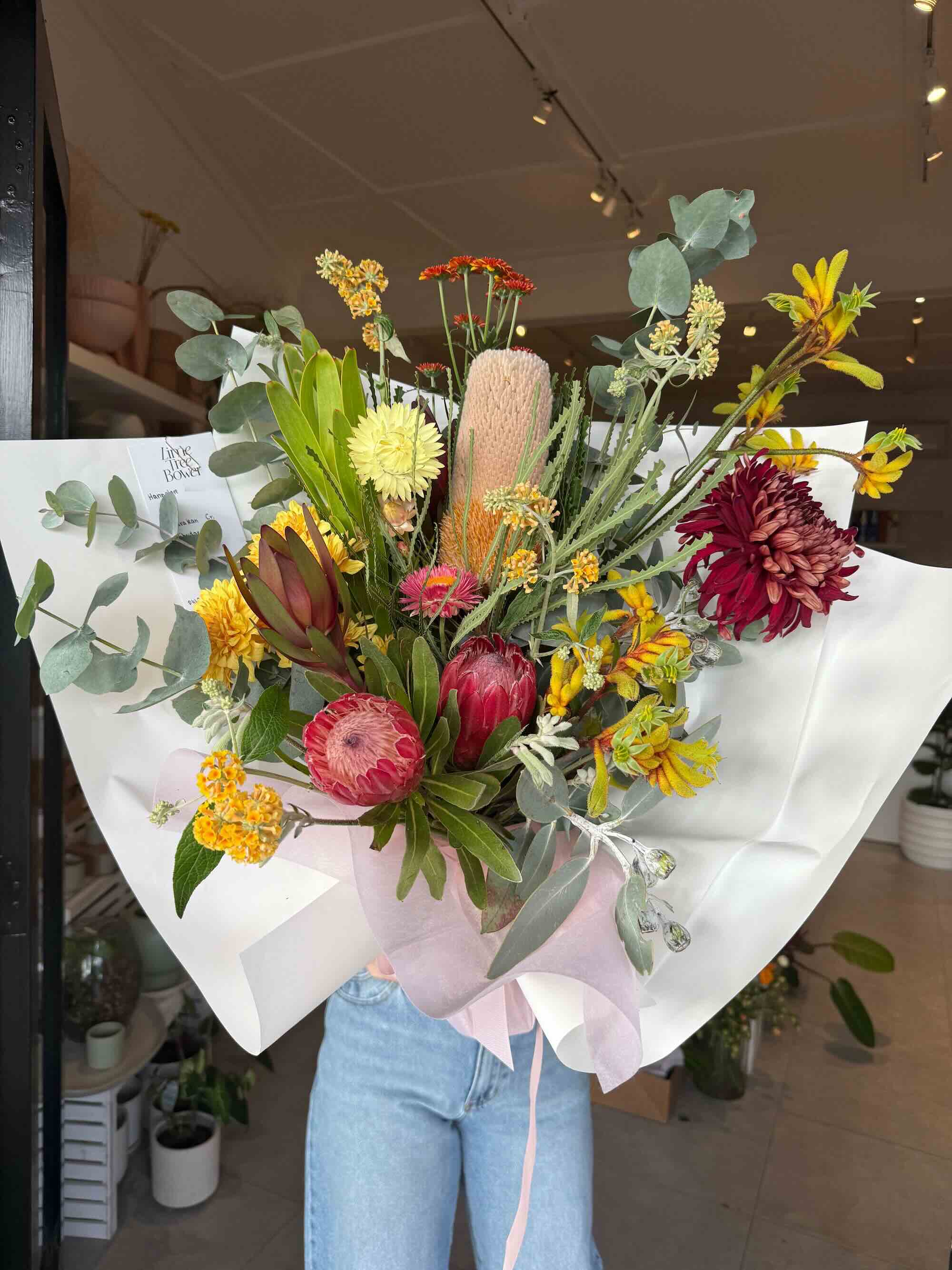 A floral mothers day natives bouquet featuring a variety of colours and textures, predominantly in earthy tones, wrapped in white paper with a pink ribbon.