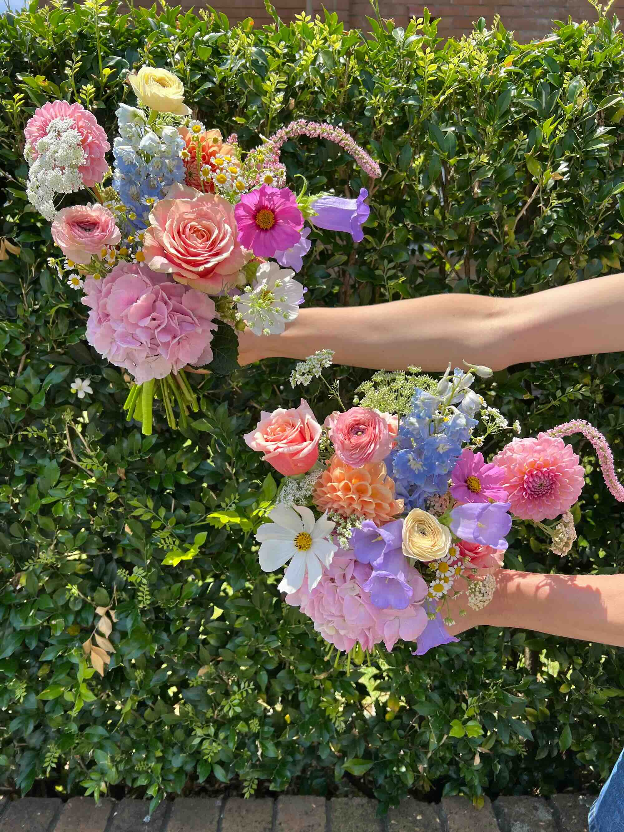Two hands holding out bridesmaid bouquets with a variety of flowers including roses and other seasonal blooms, with greenery in the background.