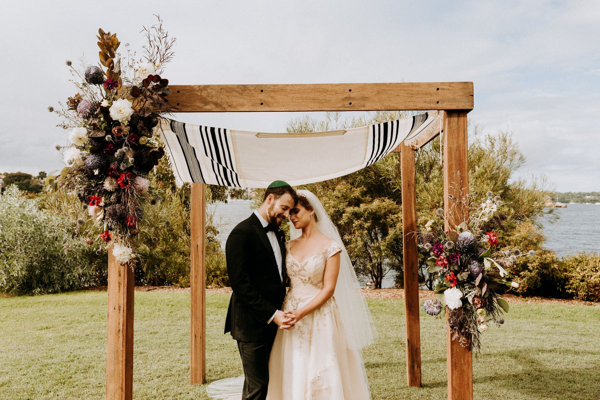Lara and Chris under a timber chuppah with a striped tallit canopy and dark flower clusters on two corners, Sydney Harbour behind