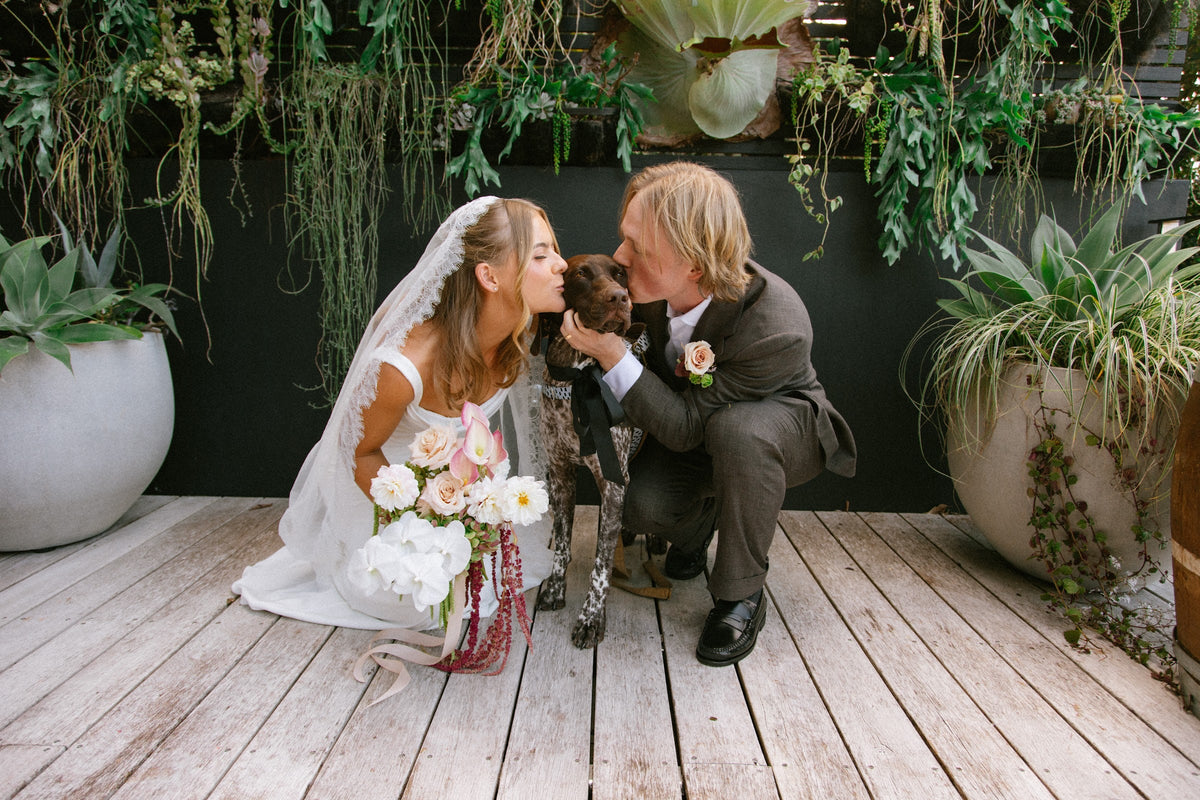 Tayla and Hamish crouching on a timber deck with their dog, her bouquet of blush roses, white dahlias and trailing amaranthus beside them