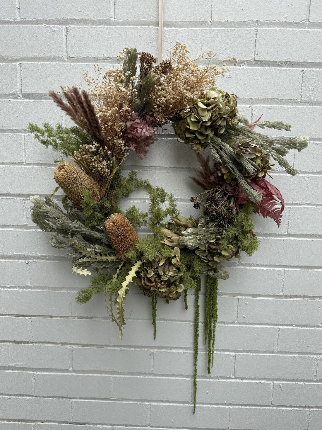A dried floral wreath with green, beige, and muted pink botanicals hanging against a white painted brick wall.