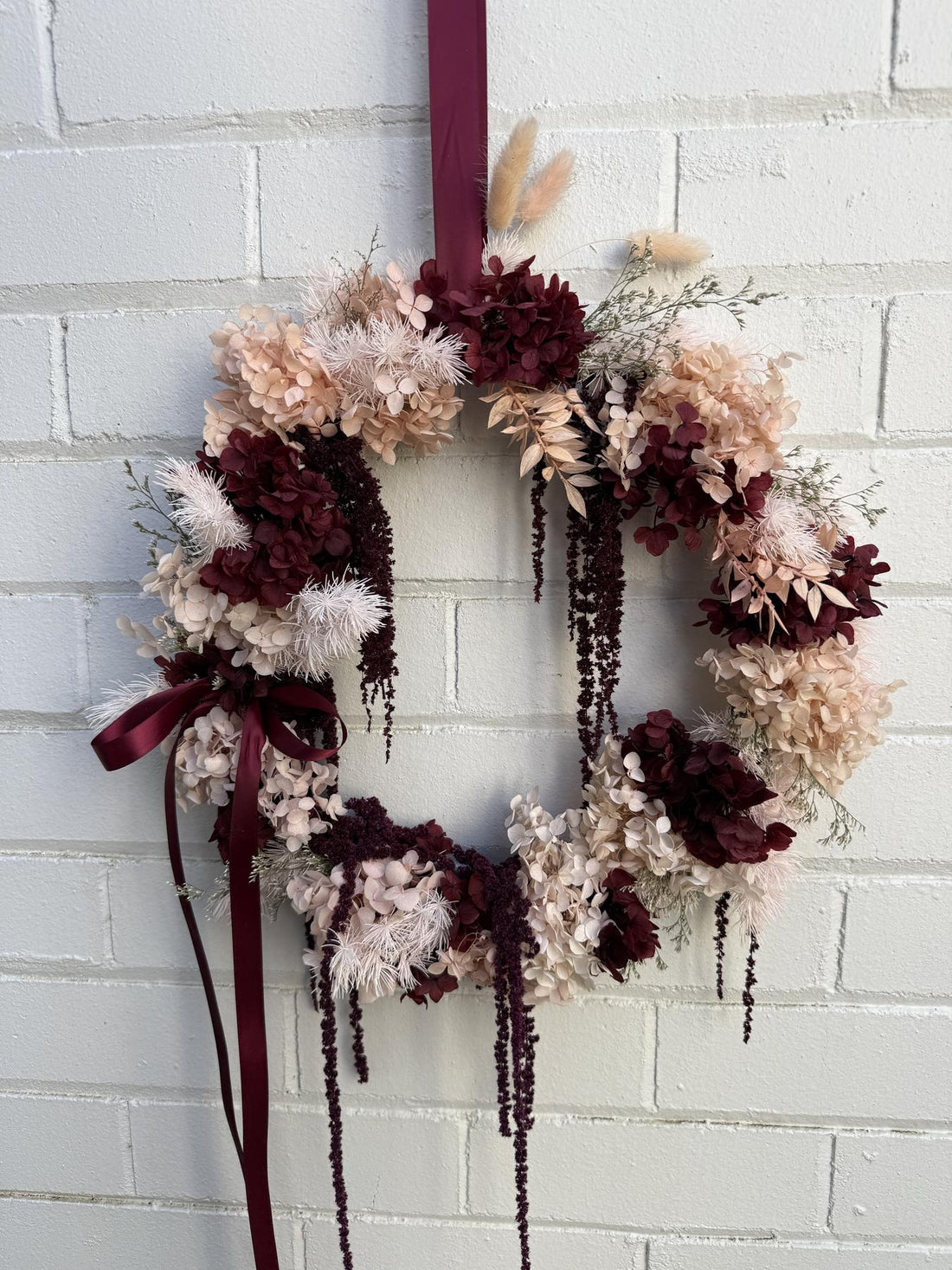 A dried floral wreath with burgundy, beige, and cream flowers, hanging on a white painted brick wall. The wreath features hanging burgundy ribbons and dried pampas grass.