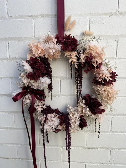 A dried floral wreath with burgundy, beige, and cream flowers, hanging on a white painted brick wall. The wreath features hanging burgundy ribbons and dried pampas grass.