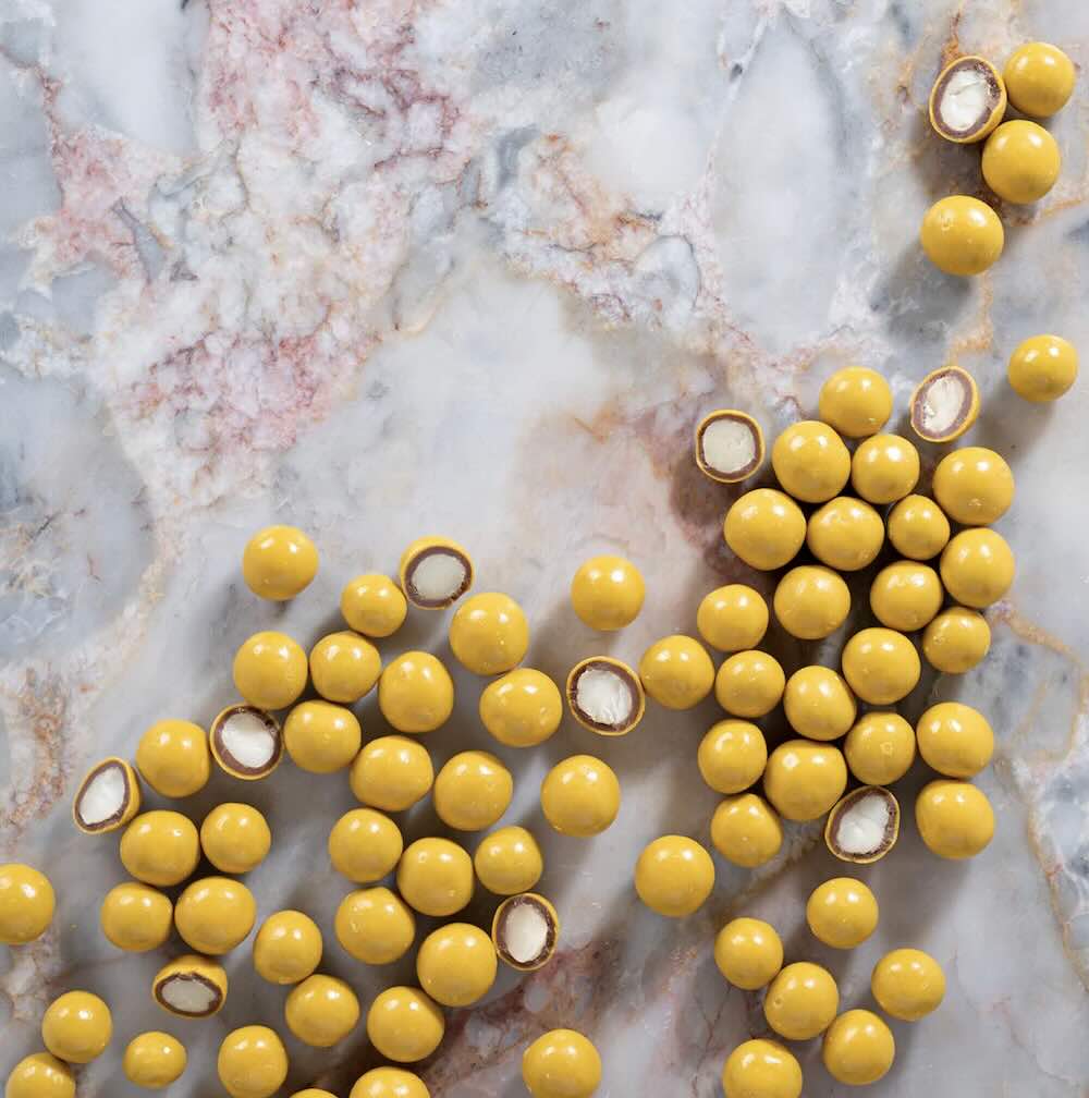 Yellow balls of milk chocolate coated passionfruit macadamia nut sweet treats scattered on a marble surface