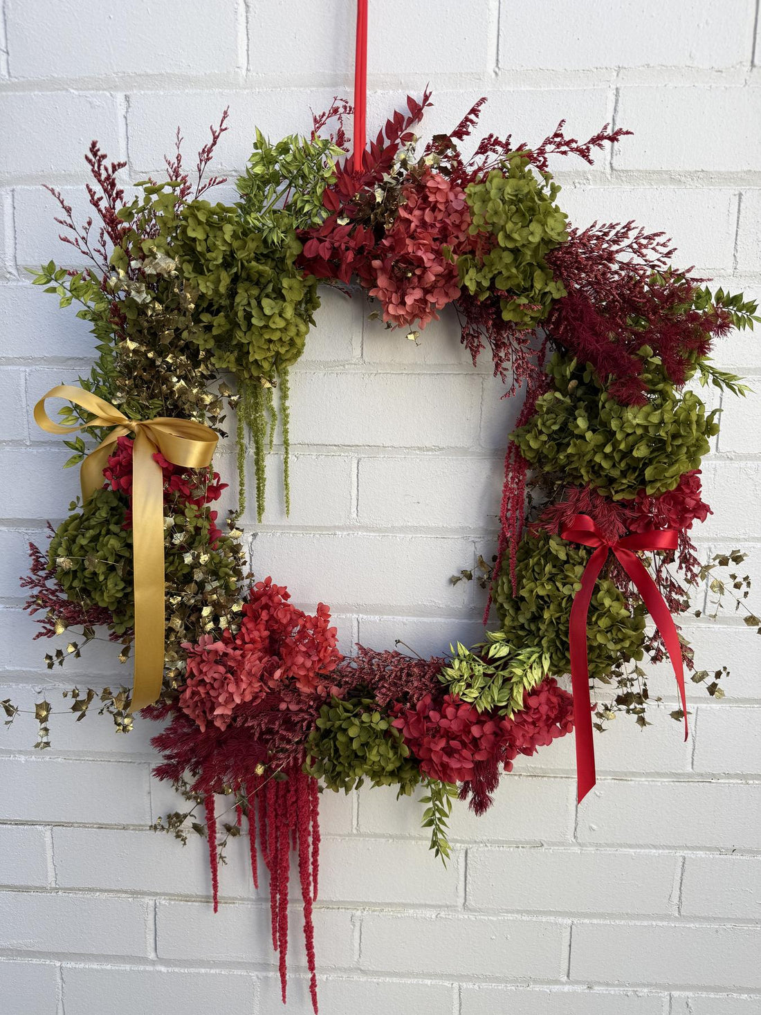A festive Christmas wreath made of green and red foliage, featuring red hydrangeas, burgundy berries, hanging red ribbons, a gold ribbon bow on the left, and a red hanging loop at the top, displayed against a white painted brick wall.