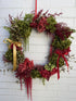 A festive Christmas wreath made of green and red foliage, featuring red hydrangeas, burgundy berries, hanging red ribbons, a gold ribbon bow on the left, and a red hanging loop at the top, displayed against a white painted brick wall.