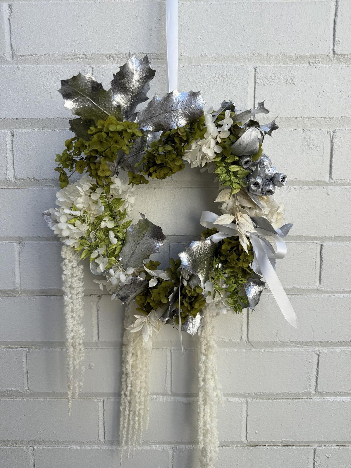 A decorative wreath hanging on a white painted brick wall. The wreath features silver faux holly leaves, green hydrangea-style foliage, white flowers, silver ribbons, and long cream-colored hanging strands.